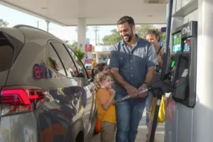 Família em posto de gasolina, abastecendo carro híbrido com etanol, refletindo economia.