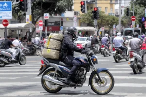 Motoboy em Honda CG 160 com capacete e mochila em semáforo vermelho cercado por outras motos em rua urbana brasileira.