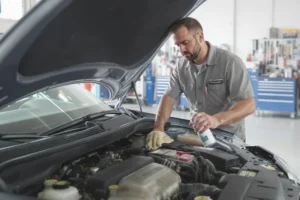 Mecânico olha para motor de carro em preparação para limpeza, segurando produtos em oficina.
