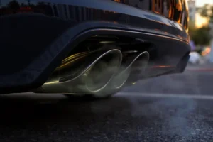Close-up de escapamento de carro esportivo metálico, sem chamas, apenas fumaça sutil, em cenário urbano ao pôr do sol.