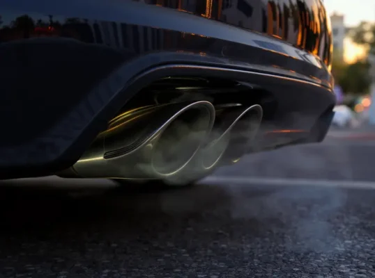 Close-up de escapamento de carro esportivo metálico, sem chamas, apenas fumaça sutil, em cenário urbano ao pôr do sol.
