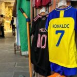 Camisa do Cristiano Ronaldo, amarela e azul, ao lado de camisa preta e rosa de Messi, em vestiário de futebol.
