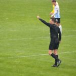 Árbitro de futebol, vestindo uniforme preto, com a mão estendida para o alto, fazendo sinal para os jogadores durante a partida.