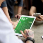 Homem de camisa social branca usando um tablet aberto em uma página de apostas esportivas verde.