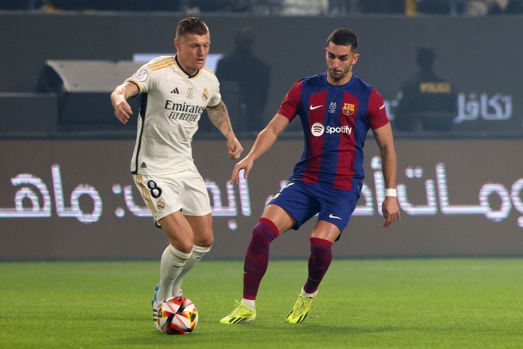 Jogador do Real Madrid com a bola dominada, enquanto jogador do Barcelona tenta realizar o desarme.