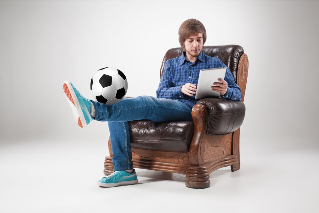 Homem de camisa azul e calça jeans, sentado em uma poltrona marrom, usando um tablet e equilibrando uma bola na canela.