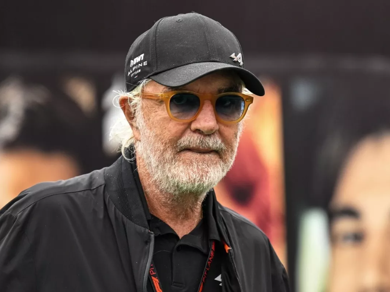 Flavio Briatore sorrindo no paddock da F1 com carros da Alpine ao fundo.