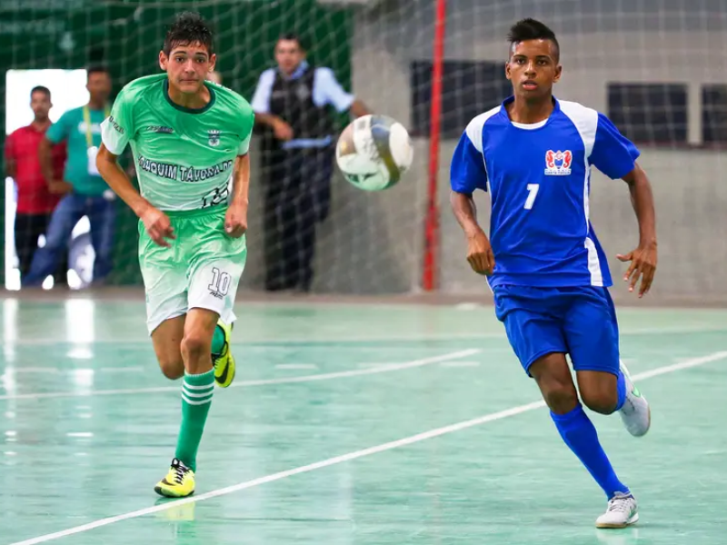Ginásio de futsal com jovens atletas em jogo, torcida vibrante e iluminação intensa, capturando a energia dos Jogos da Juventude.