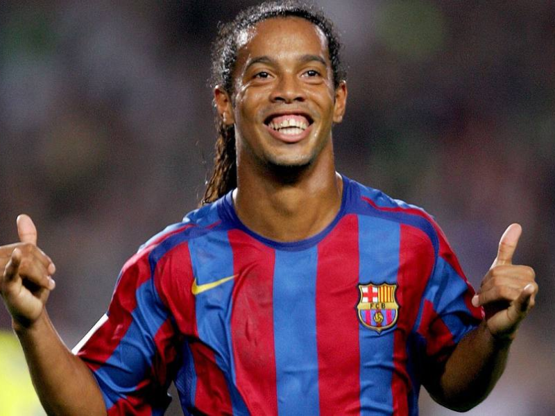 Ronaldinho Gaúcho sorrindo e driblando a bola em um jogo noturno pelo Barcelona.