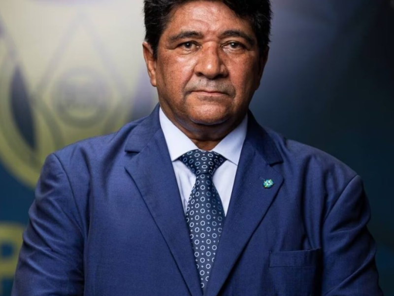 Ednaldo Rodrigues, presidente da CBF, sorrindo em seu escritório com a bandeira do Brasil ao fundo.