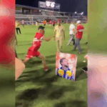 Jogador do CRB comemora o título chutando foto de rival do CSA em vestiário vibrante.