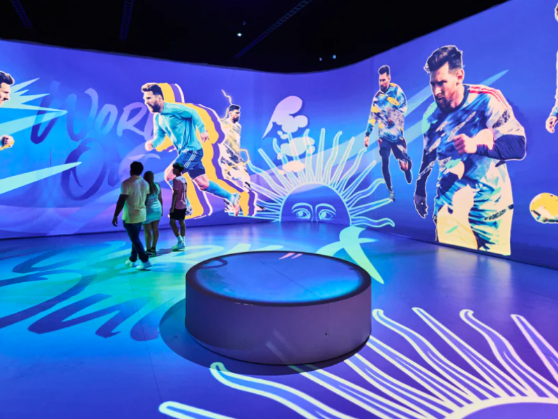 Imagem da exposição The Messi Experience em São Paulo, com fãs interagindo com hologramas de Messi.