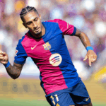 Raphinha em ação, correndo com a bola em um campo de futebol europeu.