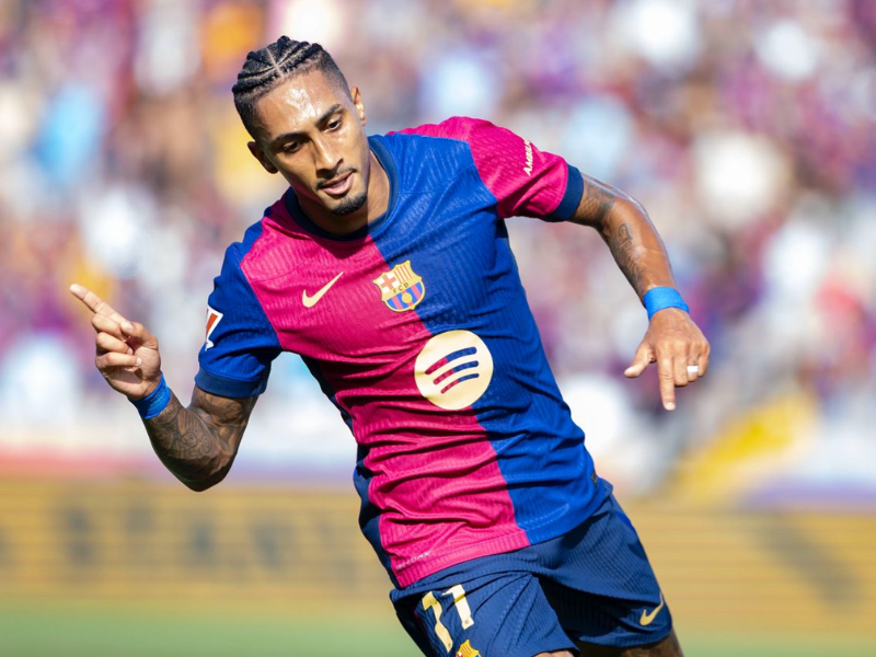 Raphinha em ação, correndo com a bola em um campo de futebol europeu.