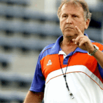 Zico em Orlando tirando uma selfie com um torcedor na Casa Flamengo, com as taças do Mundial de 1981 e Libertadores de 2022 ao fundo.