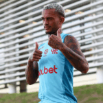 Fotografia ultrarrealista de Wesley, jogador do Flamengo, chutando a bola em campo. Ele veste uniforme vermelho e preto e a cena é iluminada por holofotes com torcedores borrados ao fundo.