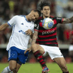 Uma fotografia ultrarrealista de uma bola de futebol perfeitamente nítida em um campo, com jogadores borrados ao fundo, representando a intensa disputa pelo título do Brasileirão entre Cruzeiro e Flamengo, sob um céu alaranjado.