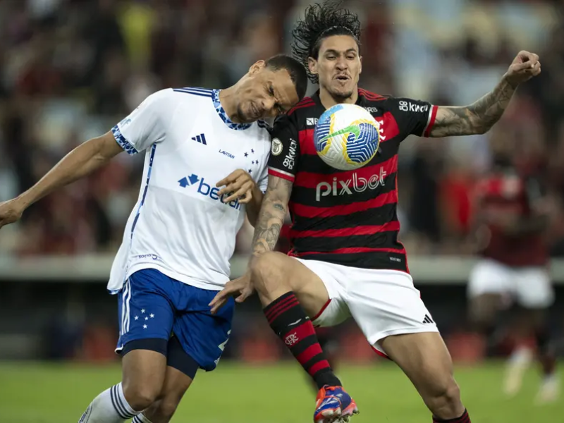 Uma fotografia ultrarrealista de uma bola de futebol perfeitamente nítida em um campo, com jogadores borrados ao fundo, representando a intensa disputa pelo título do Brasileirão entre Cruzeiro e Flamengo, sob um céu alaranjado.