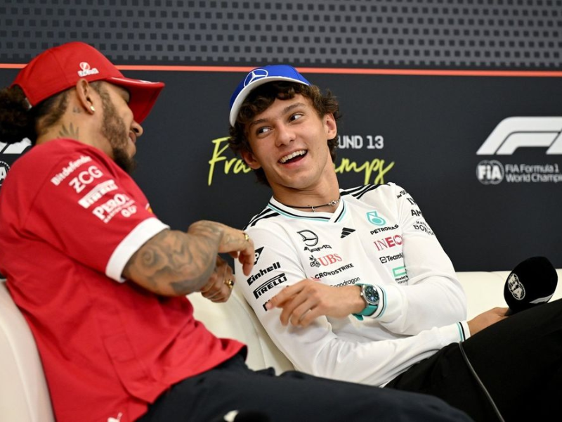 Lewis Hamilton, com camisa da Mercedes, sorri para Kimi Antonelli, que ri de forma descontraída, ambos no paddock da Fórmula 1, cercados por equipes e equipamentos.