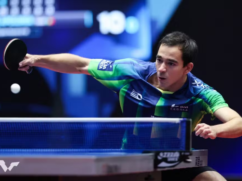 Hugo Calderano, atleta brasileiro de tênis de mesa, sorrindo e exibindo sua medalha de ouro no pescoço em uma arena de tênis de mesa. Imagem vibrante e ultrarrealista com iluminação focada.