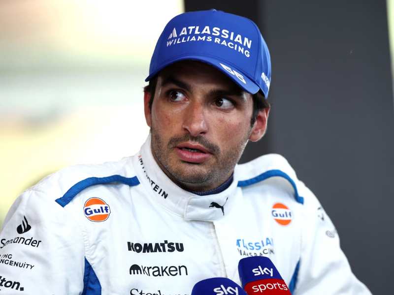 Carlos Sainz, piloto de Fórmula 1, em sua vestimenta de corrida em uma garagem, com um carro de F1 ao fundo, olhando confiante.