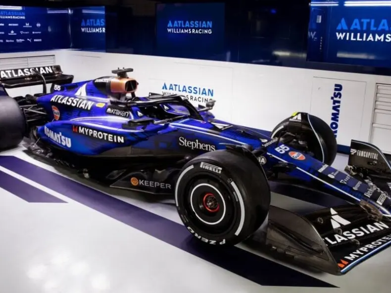 Close-up detalhado de um carro de Fórmula 1 da equipe Williams com uma pintura especial laranja e azul-escuro, exibindo palavras dos fãs. O carro está em uma pista molhada ao crepúsculo, com luzes azul e laranja acentuando os detalhes, em Interlagos São Paulo.