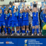 Jogadoras da Seleção Brasileira de Futsal Feminino comemorando um gol crucial contra a Colômbia, em quadra iluminada, com multidão ao fundo.