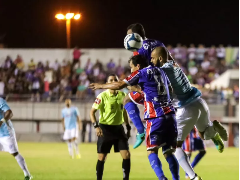 Campo de futebol iluminado à noite, com o placar ao fundo exibindo 'Itabaiana vs CSA' e '19:30'. O gramado está impecável e as arquibancadas estão repletas de uma multidão borrada, transmitindo a emoção do jogo.