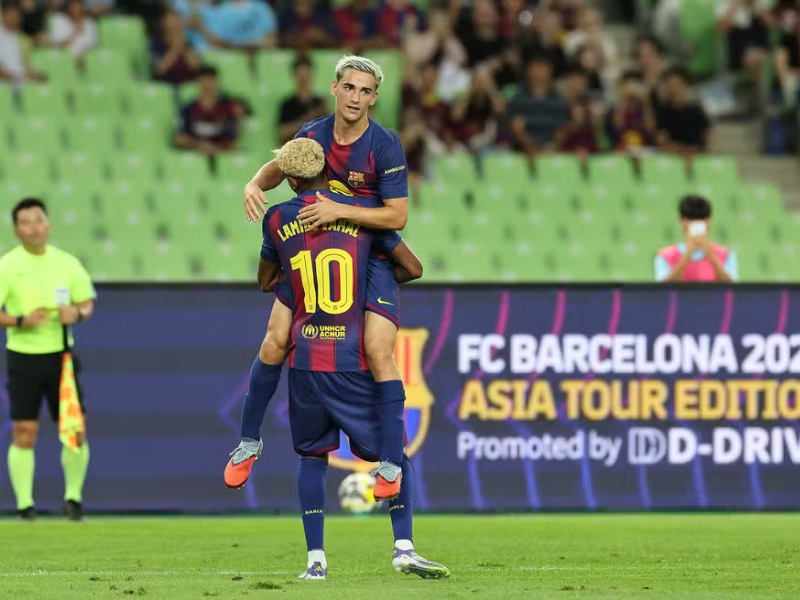 Dois jogadores do Barcelona, com uniformes vibrantes, comemoram um gol em um campo de futebol, expressando emoção e alegria.