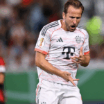 Harry Kane, jogador de futebol do Bayern de Munique, ajoelhado no gramado do estádio, comemorando um gol decisivo com os braços erguidos e expressão de euforia. A iluminação noturna realça cada detalhe de seu uniforme e suor.