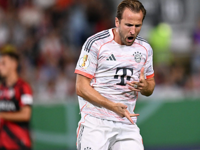 Harry Kane, jogador de futebol do Bayern de Munique, ajoelhado no gramado do estádio, comemorando um gol decisivo com os braços erguidos e expressão de euforia. A iluminação noturna realça cada detalhe de seu uniforme e suor.