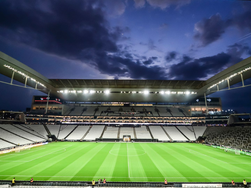 Neo Química Arena ao pôr do sol com uma mão depositando dinheiro em uma caixa de coleta para a campanha 'DOE ARENA CORINTHIANS'.
