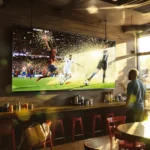 Três pessoas em um bar esportivo assistindo futebol na TV com expressões de surpresa e emoção, refletindo o clima do jogo.